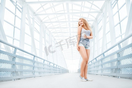 美丽的少妇有长的卷曲的头发，拿着一个带走咖啡杯和站立在反对都市背景的桥。