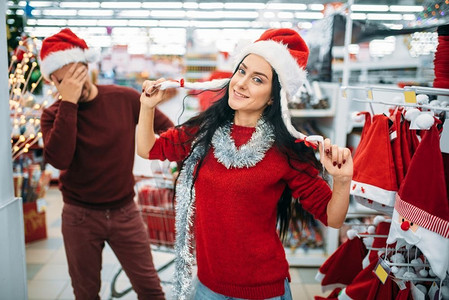 圣诞衣服摄影照片_有趣的年轻夫妇尝试圣诞衣服在超市，家庭传统。12月购物的节日商品和装饰品