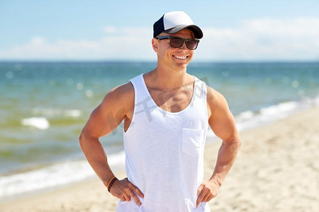 暑假和人们的概念快乐-戴着太阳镜和帽子的年轻人在海滩上微笑。戴墨镜的年轻人在夏日海滩上微笑