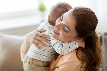 抱抱摄影照片_家庭、孩子和母亲的概念-快乐微笑的年轻母亲在家中拥抱小宝宝。快乐的年轻妈妈在家里抱着小宝宝