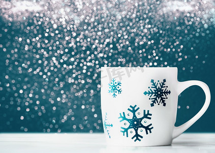 蓝色雪花图摄影照片_白色马克杯，桌子上有雪花，蓝色背景，前视概念。寒假安排