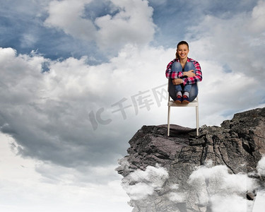 椅子创意摄影照片_站在顶端。一位穿着休闲服装的年轻女子坐在岩石上的椅子上