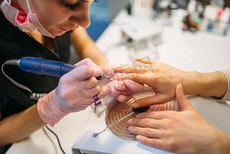 机甲人摄影照片_戴手套的美容师用抛光机在美容院治疗女顾客指甲、修指甲。美甲师做手部护理美容程序