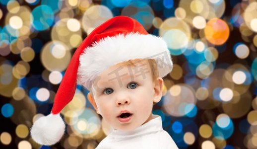圣诞节，节日和人们的概念-戴着圣诞帽的小男孩在灯光下的背景。圣诞节戴着圣诞帽的小男婴