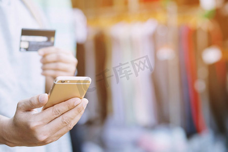 年轻男性手持信用卡和使用移动智能电话站在服装店前面。网上购物购买销售或付款。