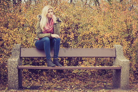悲伤的女人放松坐在公园的长凳上在秋天的天气。妇女放松坐在长凳在秋天公园
