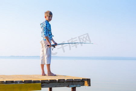 手里拿鱼摄影照片_穿着蓝色衬衫的男孩站在海边的码头上，手里拿着一根钓鱼竿