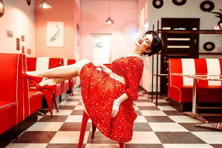 红色广告宣传摄影照片_复古咖啡馆，50美式时尚，性感化妆美女在椅子上摆姿势。红色鞋子和连衣裙，带有波尔卡圆点，复古风格