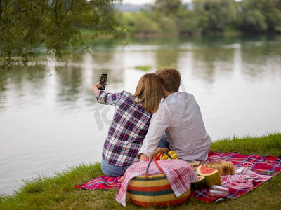 在爱的夫妇采取自拍通过手机，同时享受野餐时间饮料和食物在美丽的自然在河岸