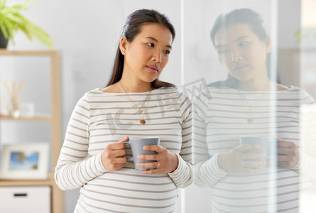 怀孕，休息，人和期望概念—悲伤的怀孕亚洲妇女与杯喝茶在家。悲伤的孕妇喝茶在家
