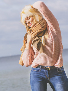 秋日时光摄影照片_在秋天的天气里，十几岁的女性在海滩上度过户外时光。穿着毛衣，戴着毛皮帽..海滩上的女人展示时尚