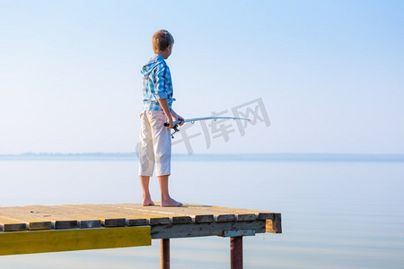 蓝色衬衫摄影照片_穿着蓝色衬衫的男孩站在海边的码头上，手里拿着钓鱼竿。穿蓝衬衫的男孩站在馅饼上