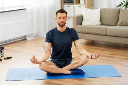 中禅摄影照片_运动，健身和健康生活方式概念—男子在家中瑜伽垫上以莲花姿势冥想。男子冥想莲花姿势在瑜伽垫在家里