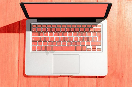 在阳光明媚的日子里，木桌上有活生生的珊瑚色键盘的笔记本电脑，从高处俯瞰。木桌上的笔记本电脑