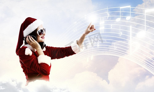 听音乐摄影照片_庆祝新年。年轻漂亮的圣诞老人女孩戴着耳机听音乐