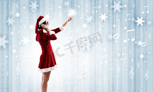 音乐圣诞老人摄影照片_庆祝新年。年轻漂亮的圣诞老人女孩戴着耳机听音乐