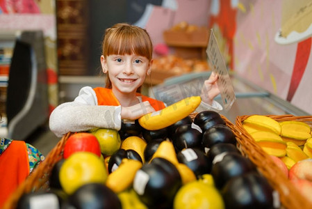 游戏室摄影照片_微笑的小女孩在制服扮演女售货员，游戏室。孩子们在想象中的超市里扮演水果销售员，销售员职业学习