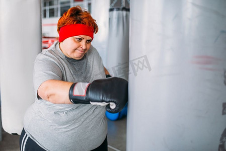 戴着拳击手套的胖女人拿着拳击袋工作，在健身房锻炼。卡路里燃烧，肥胖女性在体育俱乐部进行艰苦训练