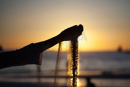 手工浇注海滩上的沙子。夕阳西下，一位年轻女孩的剪影&S手在海滩上倒沙子，映衬着黄昏的天空