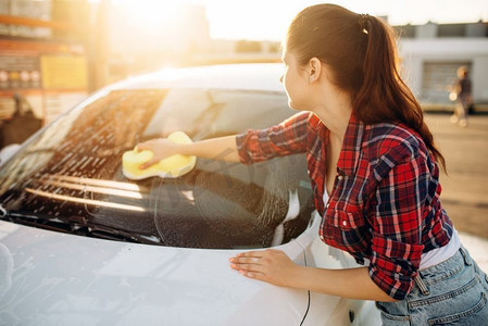年轻妇女用海绵擦洗汽车玻璃泡沫，洗车。女士自助洗车。夏日户外洗车 