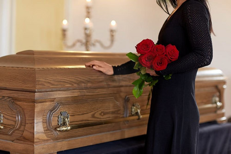 人物拿花摄影照片_葬礼，人物和哀悼的概念-教堂葬礼上戴着红玫瑰和棺材的不快乐的女人。悲伤的女人在葬礼上拿着红玫瑰和棺材