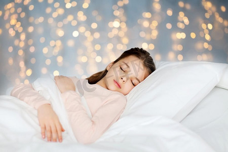 人累了摄影照片_人，孩子，休息和舒适的概念-女孩睡在床上在节日灯光背景。女孩在节日的灯光下睡在床上