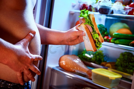 饥饿的胖子手里拿着一个大三明治，站在打开的冰箱旁边。不健康的食物。