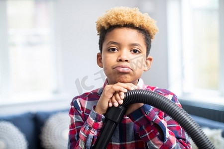 不情愿摄影照片_肖像的不快乐的男孩不情愿帮助与家务在家里拿着吸尘器