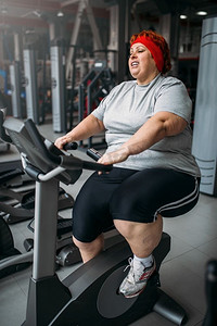 肥胖妇女训练在健身房的健身自行车。燃烧卡路里，肥胖的女性在体育俱乐部。肥胖妇女训练上健身自行车在健身房