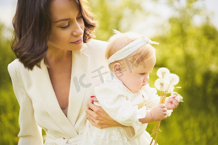 绿色春光摄影照片_年轻漂亮的母亲和可爱的小女儿在绿色的草地上吹向蒲公英的户外时尚肖像。春光映象