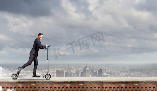 逃离办公室。年轻快乐的商人在城市背景下骑着滑板车