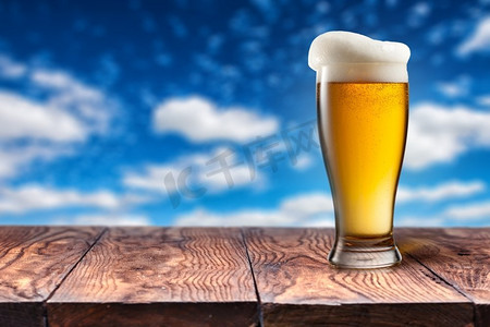 在蓝天的衬托下，木桌上放着玻璃杯啤酒，自然背景下的云朵上有波克。在蓝天的映衬下，木桌上的玻璃杯啤酒