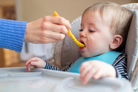 婴儿男孩在家里高椅子被喂固体食物由母亲用勺子