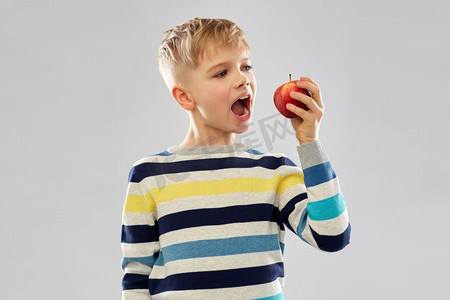 抱着东西的男孩摄影照片_食物、饮食和儿童概念--穿着条纹套头衫的男孩在灰色背景下吃红苹果。穿条纹套头衫的男孩吃红苹果