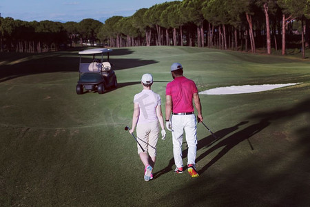 洞阳光摄影照片_一对年轻夫妇在高尔夫球场上走向下一个球洞。一个拿着高尔夫球包的男人。走在高尔夫球场上的情侣