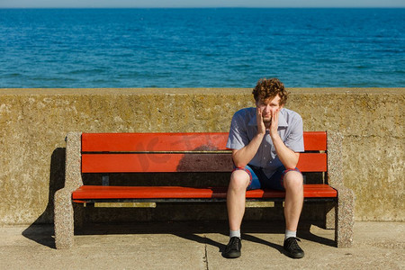 疲惫不堪的男人坐在海边的长凳上。在户外放松的年轻人。暑假..疲惫不堪的男人坐在海边的长凳上。