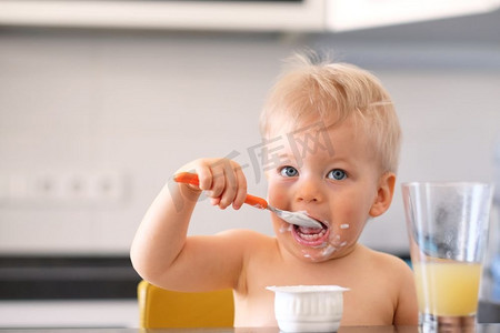 可爱的一岁男婴用勺子吃酸奶。蹒跚学步的孩子脏兮兮的脸。