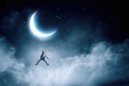 月亮上摄影照片_人挂在月亮上。一位年轻的商人在夜里吊在绳子上