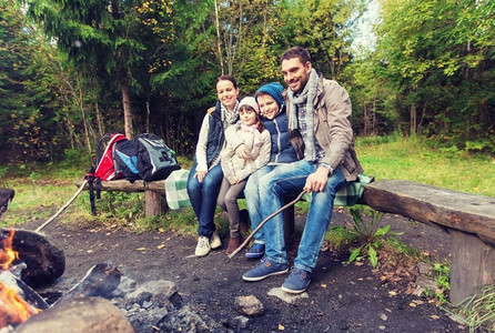 露营、旅行、旅游、徒步旅行和人的概念-坐在树林里篝火旁的长凳上的幸福家庭。幸福的一家人坐在篝火旁的长凳上