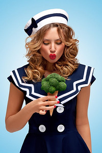 惊讶吃蔬菜摄影照片_这张极具创意的复古照片中，一位时尚的水手女孩在蓝色背景下吃着华夫饼蛋卷里的西兰花冰淇淋，看起来很惊讶。