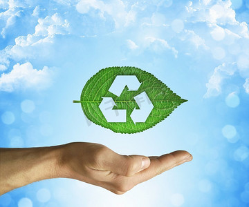 打开的手拿着一个绿色的叶子与回收的符号在蓝天背景。环境友好与可持续发展理念