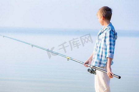 手里拿鱼摄影照片_穿蓝衬衫的男孩站在馅饼上。穿着蓝色衬衫的男孩站在海边的码头上，手里拿着一根钓鱼竿