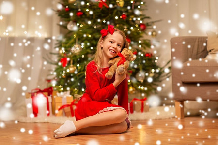 圣诞节，假日和童年概念—微笑的女孩在红色礼服拥抱泰迪熊在家。女孩在红色连衣裙拥抱泰迪熊在家里