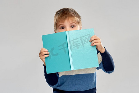 孩子的困惑摄影照片_童年和教育概念—小男孩躲在灰色背景的书后面。小男孩躲在书上