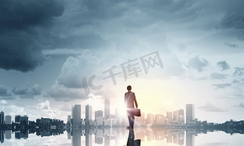 城市大景摄影照片_面对新的一天。一个商人背着背站在城市上空看着日出