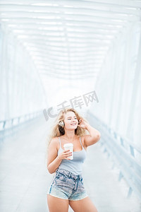 戴着音乐耳机的美丽的少妇，站在有一个带走咖啡杯和摆反对都市背景的桥