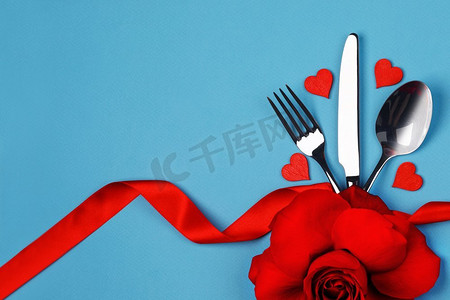 餐具套与丝带绑在蓝色背景玫瑰花和心脏情人节晚餐概念。餐具套装和心形