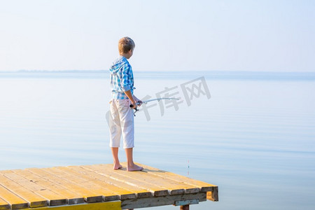 穿着蓝色衬衫的男孩站在海边的码头上，手里拿着一根钓鱼竿