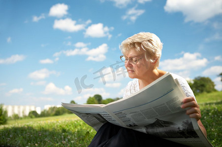 老年妇女坐在公园的草地上，在蓝天的背景下，看报纸。公园里读书