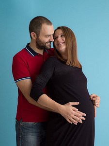 怀孕的夫妇孤立在蓝色背景。一对快乐的年轻夫妇的肖像，男子拿着他的怀孕的妻子肚子隔绝在蓝色背景
